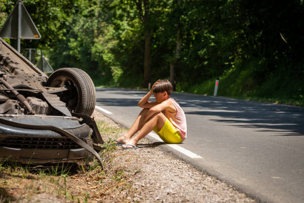 A boy in shorts and a T-shirt, a participant in a car accident, sits in shock next to an overturned car by the roadside. stock photo