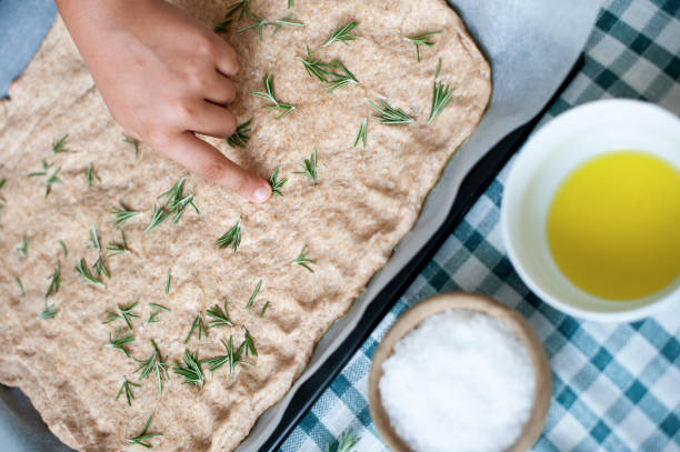 Boy Placing Rosemary Leaves over Focaccia Dough in Baking Tray stock photo