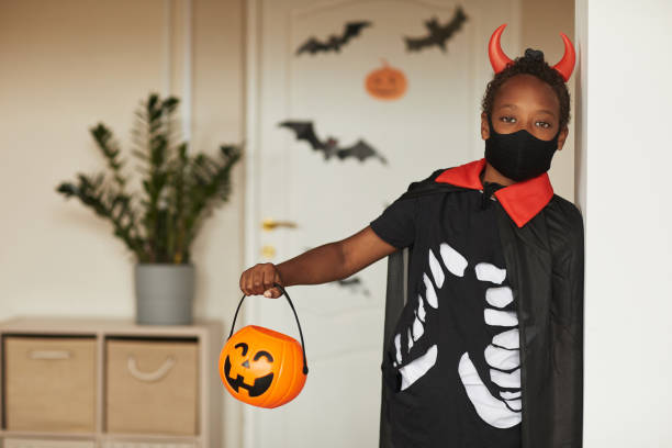 Boy Ready To Trick Or Treat Portrait of cute African American boy wearing spooky devil costume with red horns and mask holding jack o' lantern basket for candies looking at camera stage costume stock pictures, royalty-free photos & images