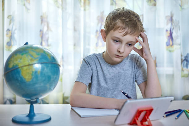 Boy thinking while looking at tablet while doing homework at home stock photo