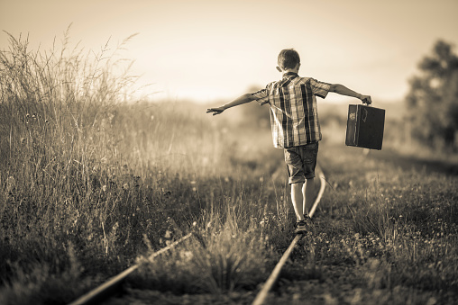 Rear view of a boy holding a suitcase in his outstretched arms. His is balancing jauntily on rails in the evening sun. Selective focus on the boy, photo is sepia toned and vignette added for retro style.