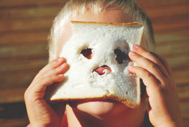 A young child holds a piece of chewed bread with a face chewed out of it obscuring his own face.