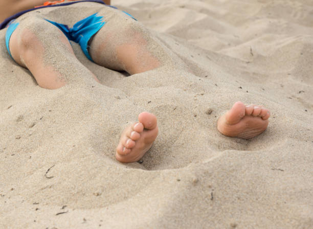 boy with half-buried feet in the sand of the beach boy with half-buried feet in the sand of the beach with blue swimsuit human feet buried in sand. summer beach stock pictures, royalty-free photos & images