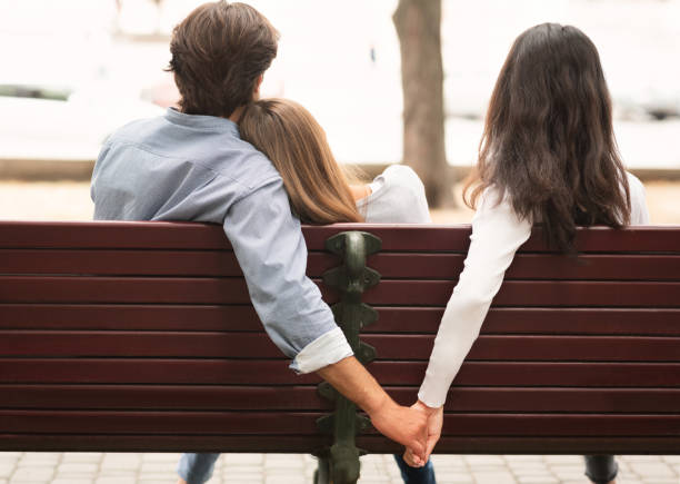 Boyfriend Holding Hands With Girlfriend's Friend Sitting On Bench Outdoor Love Triangle. Cheating Boyfriend Hugging Girlfriend Holding Hands With Her Girl Friend Sitting On Bench Together In Park Outdoor. Back-View betrayed stock pictures, royalty-free photos & images