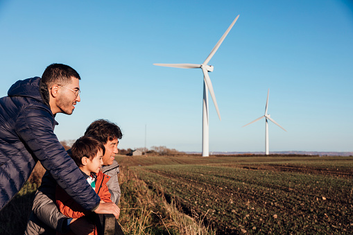 An Asian grandfather, father and son leaning against a fence in a field together, they are looking at the view together and there are wind turbines in the distance.