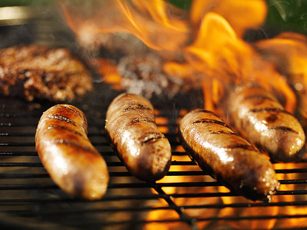 bratwursts cooking over fire on barbecue grill bratwursts cooking over fire on barbecue grill with hamburgers in background with flames. sausage stock pictures, royalty-free photos & images