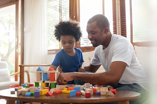 Brazilian Father and his Little son playing building tower from wooden block toy on sofa in Living room. Happy African American Man and boy enjoying at home. Leisure Activities on Holidays