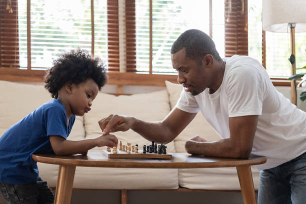 Brazilian Father and little boy playing chess on table at home together. Happy Black African American Family engaged in board game on holiday. stock photo