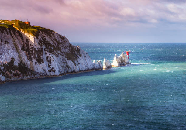 A break in the clouds illuminates the iconic chalk stone pinnacles of The Needles and the 19th century lighthouse on the coastline Isle of Wight an island off the south coast of England stock photo