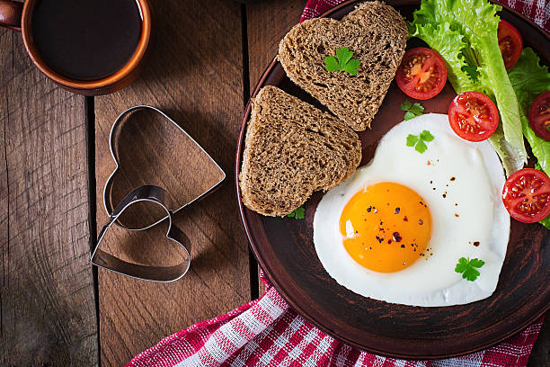 Breakfast on Valentine's Day - fried eggs Breakfast on Valentine's Day - fried eggs and bread in the shape of a heart and fresh vegetables. Top view fried egg photos stock pictures, royalty-free photos & images