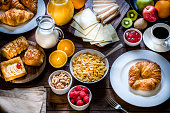 High angle view of a wooden table full of breakfast food like croissants, corn flakes, a coffee cup, marmalade, some fruits, an orange juice and a milk jugs, ham, cheese and some desserts. This image is ideal for breakfast and/or a buffet concept. Low key DSLR photo taken with Canon EOS 6D Mark II and Canon EF 24-105 mm f/4L