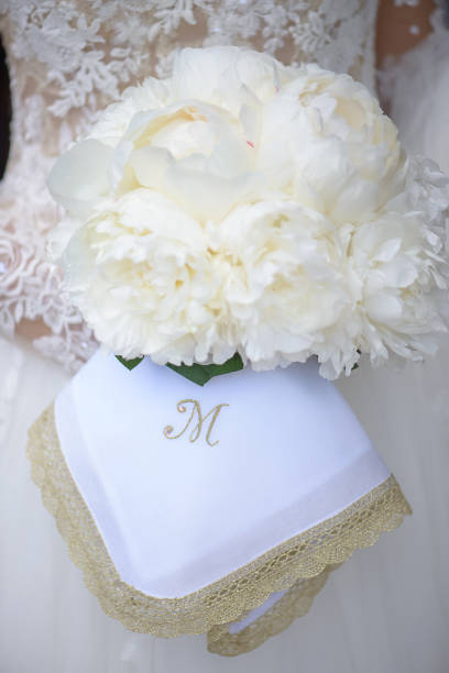 Bride holding an exquisite white peonies wedding bouquet and an embroidered linnen napkin monogrammed with letter M stock photo