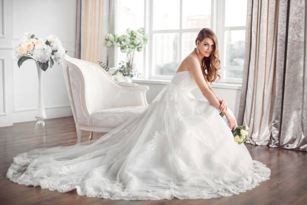 Bride in beautiful dress sitting resting on sofa indoors Wedding. Bride in beautiful dress sitting on sofa indoors in white studio interior like at home. Trendy wedding style shot in full length. Young attractive caucasian brunette model like a bride against big window tender posing. wedding dress stock pictures, royalty-free photos & images