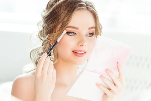 Bride Wedding Makeup. Bridal Make up. Beautiful Woman looking at Hand Mirror and putting Eye Shadow Eyebrow Self Make up in White Room stock photo