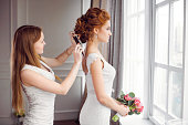 Master stylist makes the bride wedding hairstyle using hairbrush indoors at home near window. Beautiful bride perfect style. Wedding hairstyle make-up luxury wedding dress.
