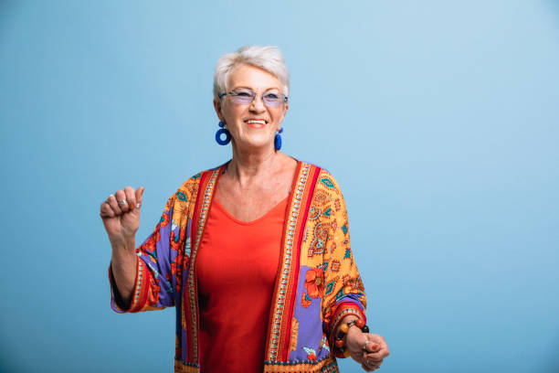 Portrait of a smiling senior woman dancing in front of a blue background. She is dressed in bright clothing and is smiling, looking away from the camera.