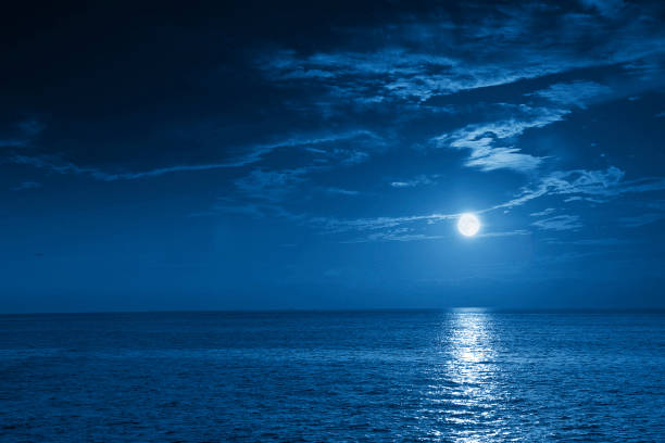 Bright Full Blue Moon Rises Over A Calm Ocean View This photo illustration of a deep blue moonlit ocean at night with calm waves would make a great travel background for any coastal region or vacation, emphasizing the beauty of the night time ocean or sea. moonlight stock pictures, royalty-free photos & images