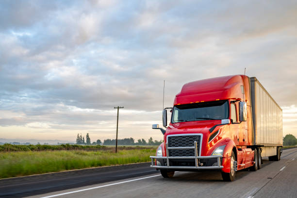 Bright red big rig bonnet semi truck transporting cargo in dry van semi trailer running on the evening highway road stock photo