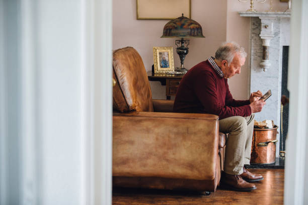 Bringing Back Memories Senior man is sitting in an armchair in the living room of his home, holding and looking at an old photo with a sad expression. armchair photos stock pictures, royalty-free photos & images