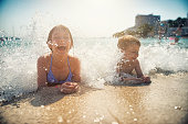 Little boy and littel girl are having fun lying on beach and being splashed by waves. Candid smile and laughter. Kids are aged 9 and 5.