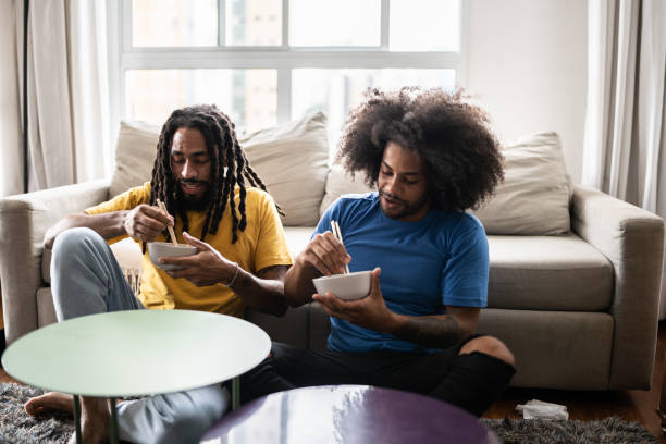 frères assis par terre en train de manger des nouilles à la maison - african america man eating chinese food photos et images de collection