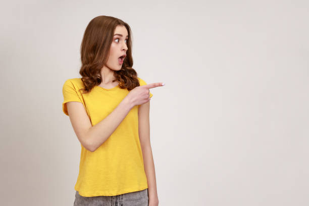 Brown haired woman of young age in yellow casual T-shirt pointing away looking with big eyes and open mouth, surprised with advertising, empty space. stock photo