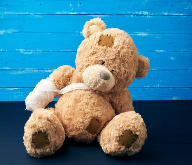 brown teddy bear with rewound white bandage paw on a blue wooden background brown teddy bear with rewound white bandage paw on a blue wooden background, pediatrics concept broken doll 1 stock pictures, royalty-free photos & images