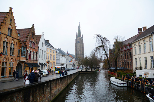 Bruges, Belgium - December 27, 2021: View of the Bruges canal with the Church of Our Lady in the background. Tourists and boats in the foreground.