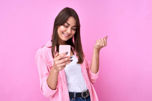 Brunette attractive girl holds mobile phone in her hand and rejoices at the good news, clenches her fists isolated in pink studio with copy space stock photo
