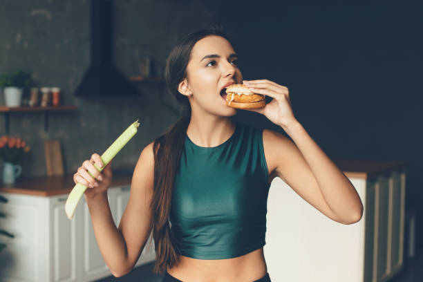 Brunette sporty woman is eating a burger while holding a leek in her hands stock photo