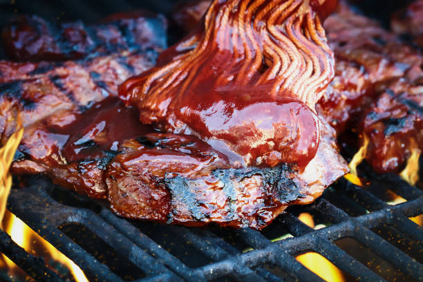 BBQ brush over boneless beef ribs grilling over flames Boneless beef ribs grilling over flames with barbecue sauce added with bbq mop. Extreme shallow depth of field with blurred background with focus on front meat. Grilled Boneless Beef Short Ribs stock pictures, royalty-free photos & images