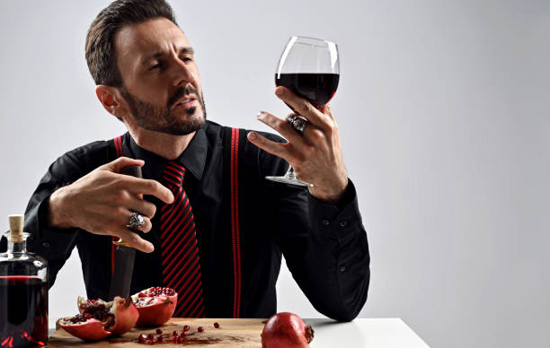 Brutal man in black shirt with striped tie and suspenders sits at table with pomegranate and juice, holds glass in hand Stylish brutal man in black shirt with striped tie and suspenders sits at table served with fresh pomegranate and juice, holding glass in hand as if he is a sommelier, expert tasting drink snob stock pictures, royalty-free photos & images