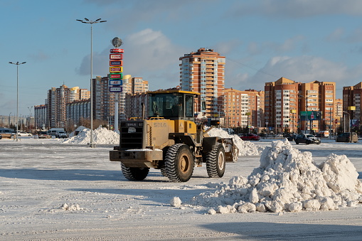 Krasnoyarsk, Krasnoyarsk Territory, RF - November 26, 2021: A bucket loader removes snow from snowdrifts in a city square after a snowfall against the backdrop of residential buildings on a sunny day.