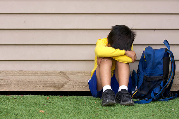 Bullied young boy sitting on grass with his head in his lap stock photo