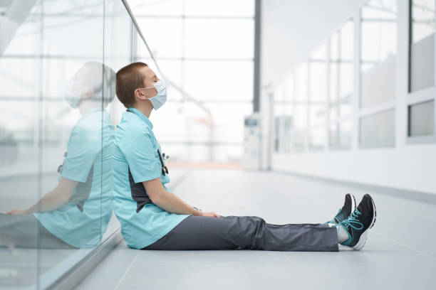 Burnout concept. Exhausted caucasian female short haircut doctor, wearing medical mask, sitting on a floor in corridor of clinic with closed eyes Burnout concept. Exhausted caucasian female short haircut doctor, wearing medical mask, sitting on a floor in corridor of clinic with closed eyes mental burnout stock pictures, royalty-free photos & images