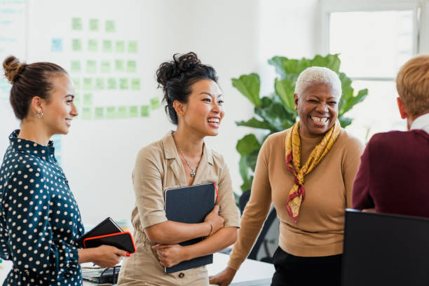 Business Can be Fun Colleagues standing in a small group discussing something while laughing. Two of the women are holding notebooks. diversity and inclusion stock pictures, royalty-free photos & images