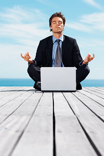 Middle aged business executive meditating on pier while laptop in...