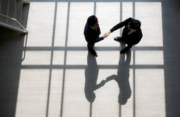 Business partners shaking hands in the modern office. Standing near the window while the sunlight shines on them. There is a shadow on the wooden floor stock photo