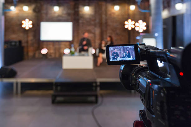 Business people during video conference Businesswoman and businessman giving presentation during online seminar, sitting on armchairs on the stage, holding microphones. Focus on video camera on the foreground. recorded webinar stock pictures, royalty-free photos & images