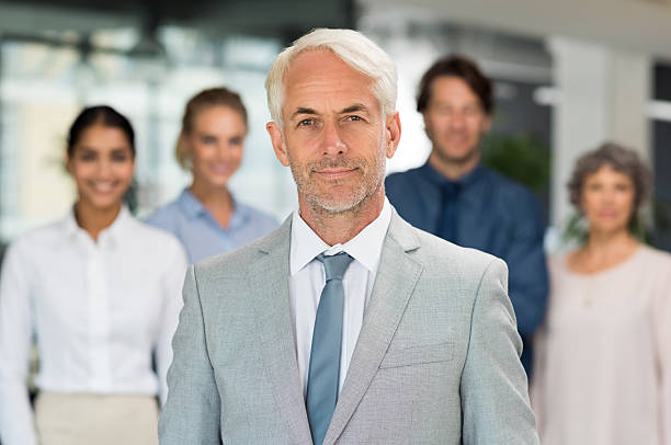 Business team Successful mature businessman standing with businesspeople behind. Portrait of senior manager looking at camera in front of his business team. Happy smiling executive in suit. cfo stock pictures, royalty-free photos & images