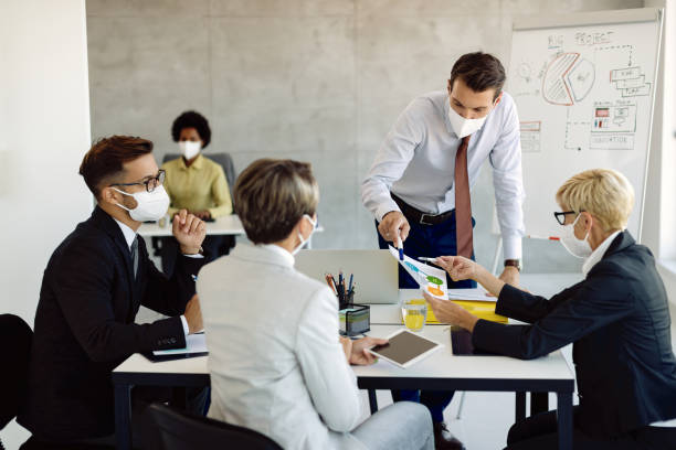 Business team with face masks cooperating while examining charts in the office. Group of entrepreneurs with protective face masks brainstorming while analyzing business graphs on a meeting in the office. social responsibility stock pictures, royalty-free photos & images