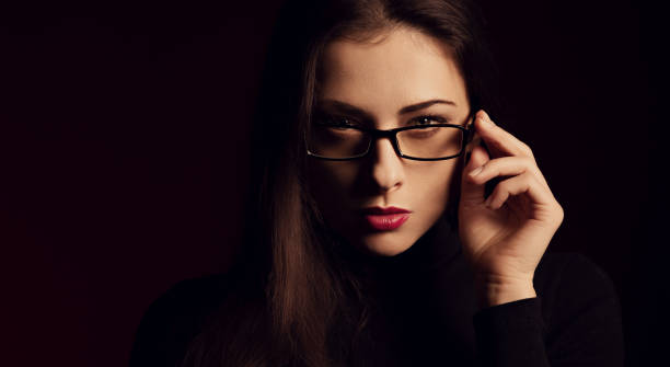 Business thinking woman in fashion eye glasses looking with hand near the face in black t-shirt on dark shadow red background. Closeup stock photo