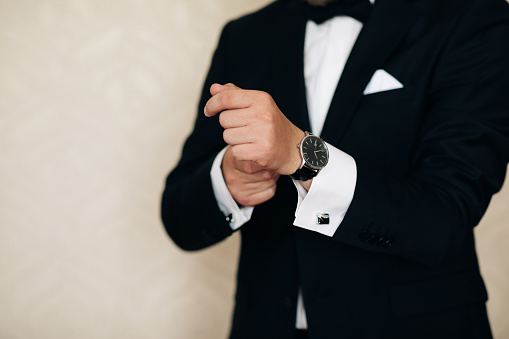 Businessman adjusts his white shirt button. Concept successful businessman. Portrait of trendy attractive stunning man in black tuxedo with tie fasten button on sleeve cuffs of his white shirt