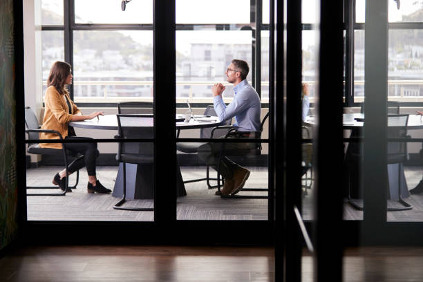een zakenman en jonge vrouw vergadering voor een sollicitatiegesprek, volledige lengte, gezien door glazen wand - interview stockfoto's en -beelden