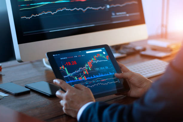 デジタルタブレットで株式市場をチェックするビジネスマンと画面上の証券取引所のグラフを持つデスクトップコンピュータ。金融市場。オフィスバックグラウンドでのデータ分析 - 株 ストックフォトと画像