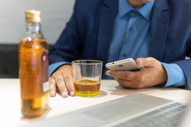 businessman drinks alcohol and looks at information through his smartphone while sitting at a table next to a laptop in a bar. Drink alcohol in the workplace. Alcoholic employee stock photo