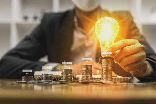 A businessman holds a glowing light bulb on top of the highest pile of coins. Placing coins in a row from low to high is comparable to saving money to grow more. Savings growth concept. stock photo