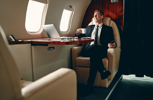 Senior businessman in suit flying in private jet and listening to music via wireless earphones.