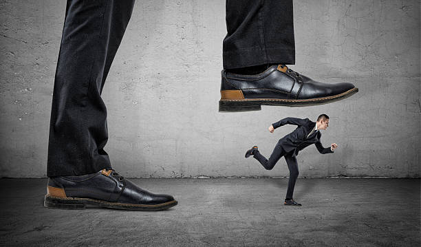 Businessman is running away from big foot on gray concrete A businessman is running away from a big foot on a gray concrete background. Suppression of competitors. Big business. broken suitcase stock pictures, royalty-free photos & images