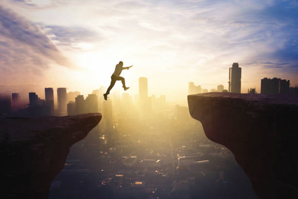 Businessman jumping through gap cliff at sunrise Young businessman jumping through gap cliff at sunrise time with modern city background courage stock pictures, royalty-free photos & images